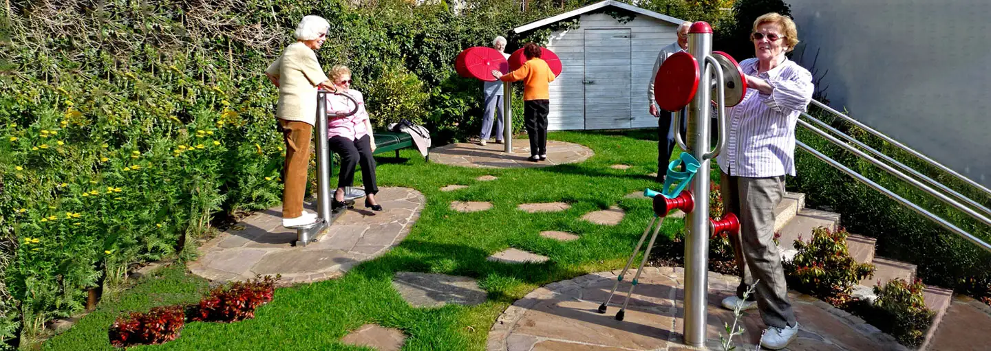 Senioren trainieren an Fitnessgeräten in einem Garten mit grünem Rasen und Büschen.