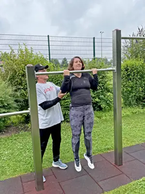 Foto einer jungen Frau die an der Reckstange versucht einen Klimmzug zu machen, rechts von ihr steht ein Herr der sie leicht von der Seite stützt.