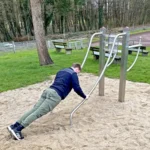 Foto des Beinheber-Liegestütz-Kombitrainers, er steht auf einer feinen Sandfläche und ein Mann führt darauf die Übung Liegestütze aus.