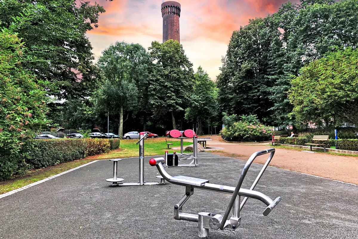 Fitnessgeräte im Freien, leeres, parkähnliches Umfeld mit Bäumen, Himmelturm im Hintergrund, Sonnenuntergang.