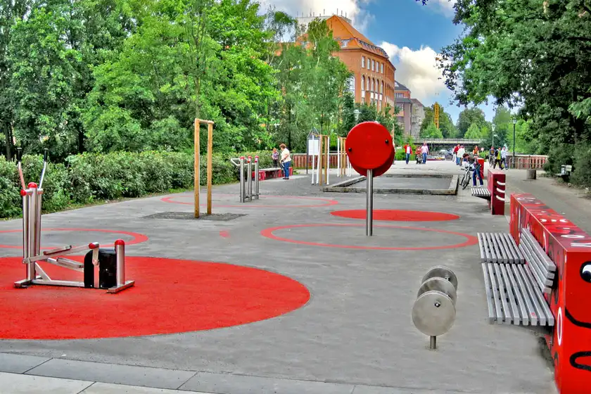 Fitnessgeräte im Freien auf städtischem Spielplatz, im Hintergrund Bäume und Gebäude, Menschen spazieren und radeln.