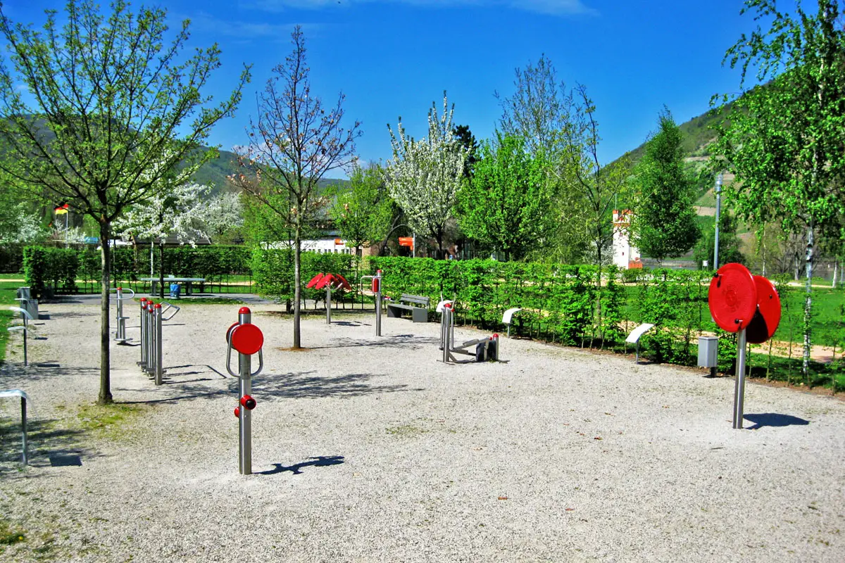 Fitnessgeräte im Freien auf Kiesweg, umgeben von grünen Bäumen und sonnigem Himmel im Park.