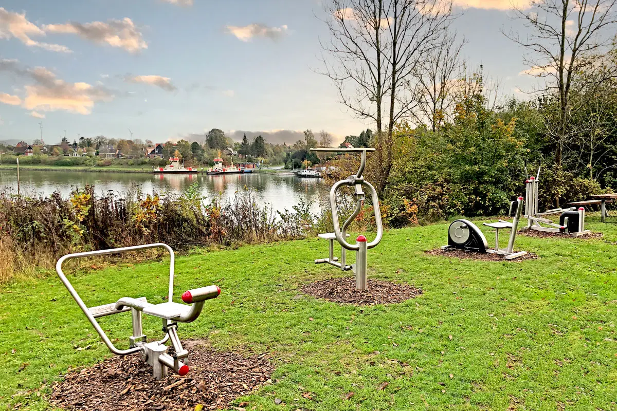 Outdoor-Fitnessgeräte auf einer Rasenfläche, mit Blick auf einen Fluss und das gegenüberliegende Ufer mit Bäumen und Gebäuden.